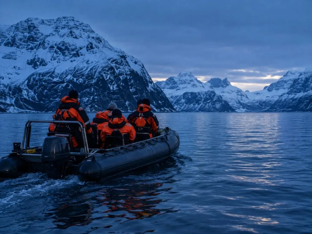 Des passagers en combinaisons thermiques agrippés aux poignées d’un RIB ouvert traversant un fjord sombre.
